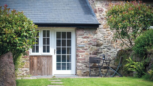 Ein Steinhaus mit weißer Tür, ein kleiner Garten mit Rasen und trittfesten Steinen, sowie ein Metallstuhl.