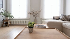 Een moderne woonkamer met een houten vloer, een beige bank, een salontafel, een potplant, een radiator en een raam met dunne gordijnen.