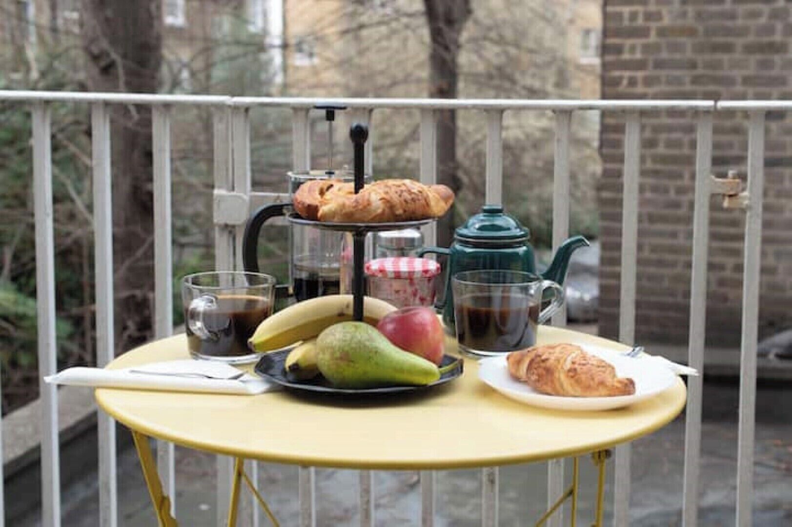 A table with a teapot, cups, a plate with fruits, and pastries on a balcony.