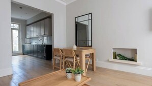 Een moderne woonkamer met een houten eettafel, twee stoelen, een klein bijzettafeltje en een potplant. Een grote spiegel hangt aan de muur, en een in de muur verwerkte planken met planten is zichtbaar.