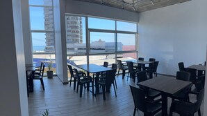 A modern dining area with multiple tables and chairs, large windows offering a view of the cityscape, and a concrete ceiling with recessed lighting. - Vistara Suites (Iquique)