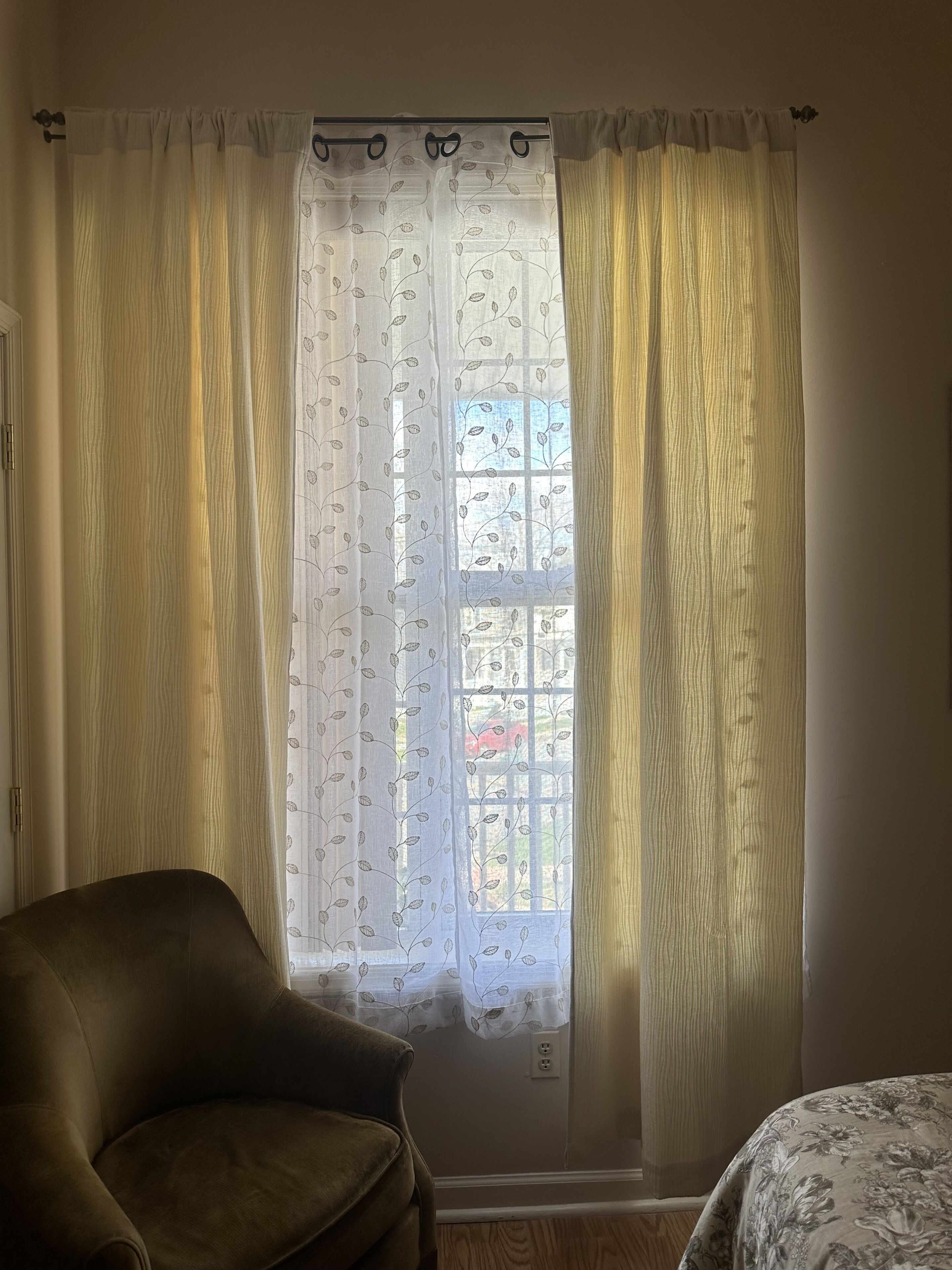 A room with a window covered by sheer curtains with a leaf pattern, a brown leather chair, and a bed with a floral bedspread.