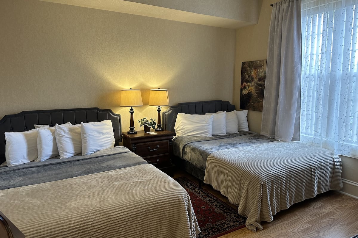 A hotel room with two beds, a nightstand with lamps, a painting on the wall, and a window with sheer curtains.