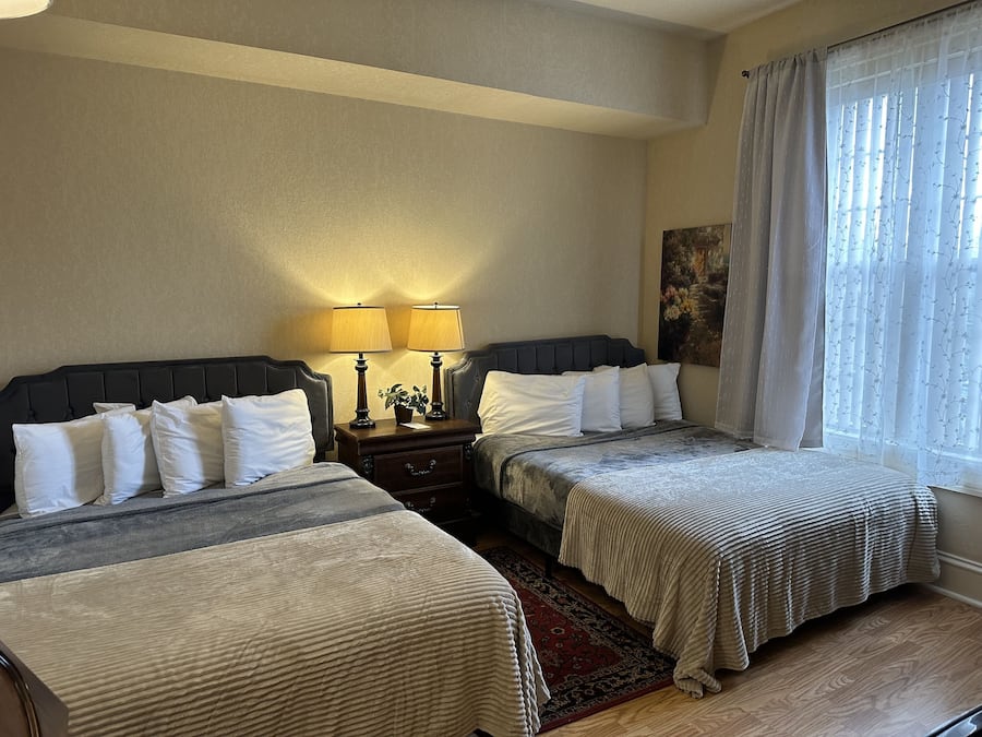 A hotel room with two beds, a nightstand with lamps, a painting on the wall, and a window with sheer curtains.