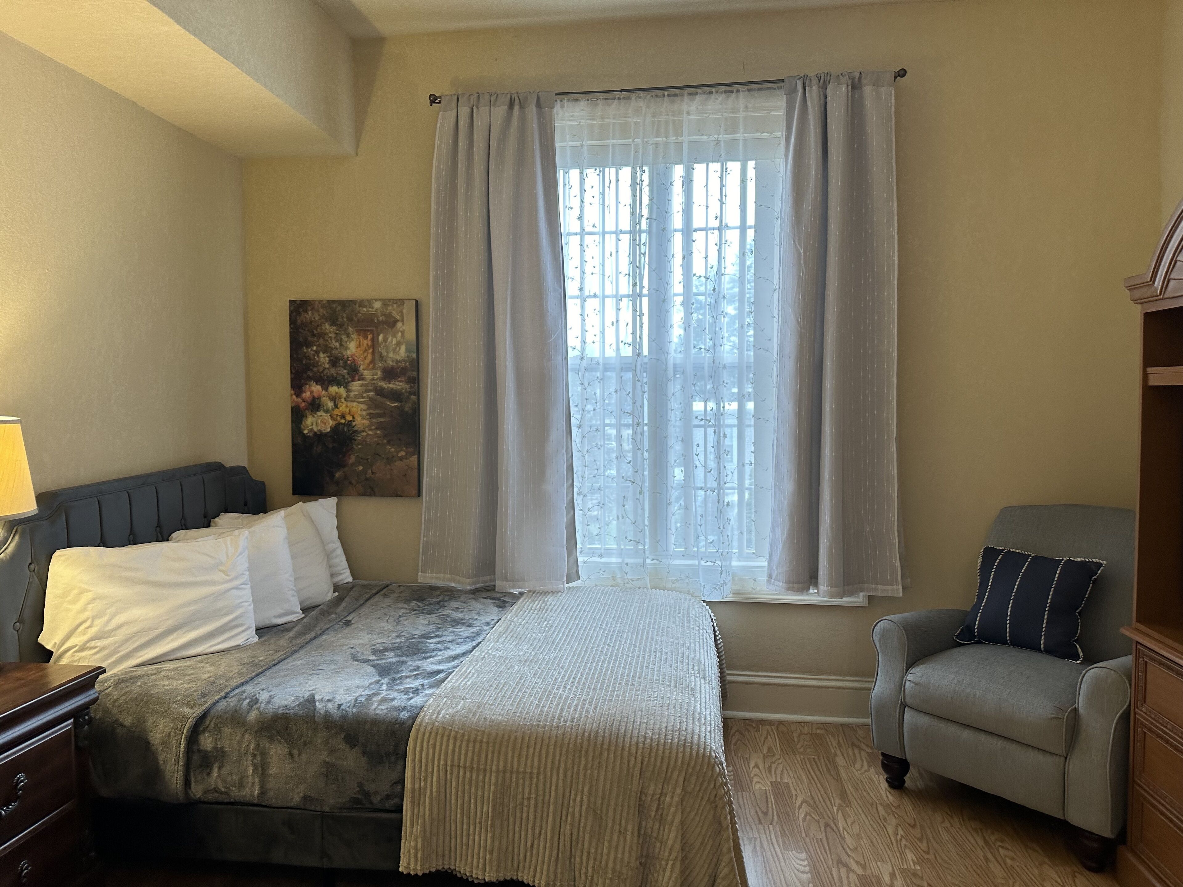 A bedroom with a bed, a chair, a window with curtains, a painting on the wall, and a lamp.