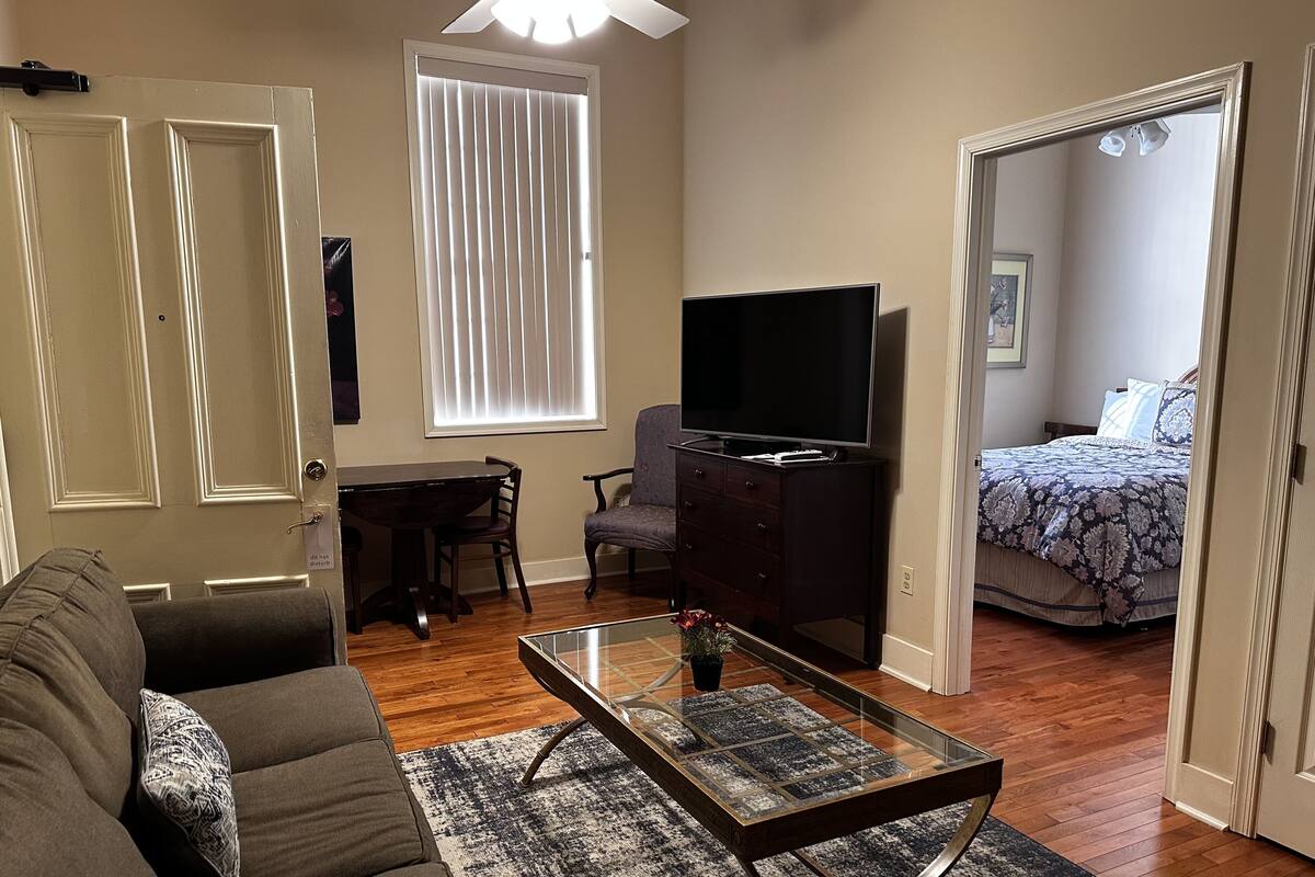 A living room with a sofa, a glass coffee table, a TV on a stand, a dining table with chairs, and a bedroom with a bed and a window.