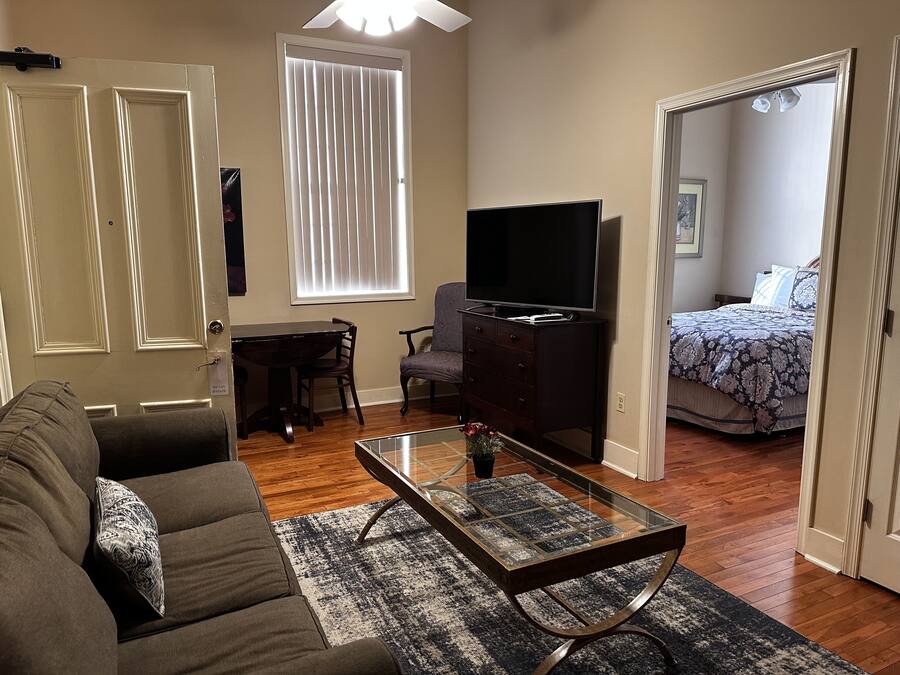 Una sala de estar con sofá, una mesa de centro de vidrio, un televisor sobre un mueble bajo, una mesa de comedor con sillas, y un dormitorio con cama y ventana.