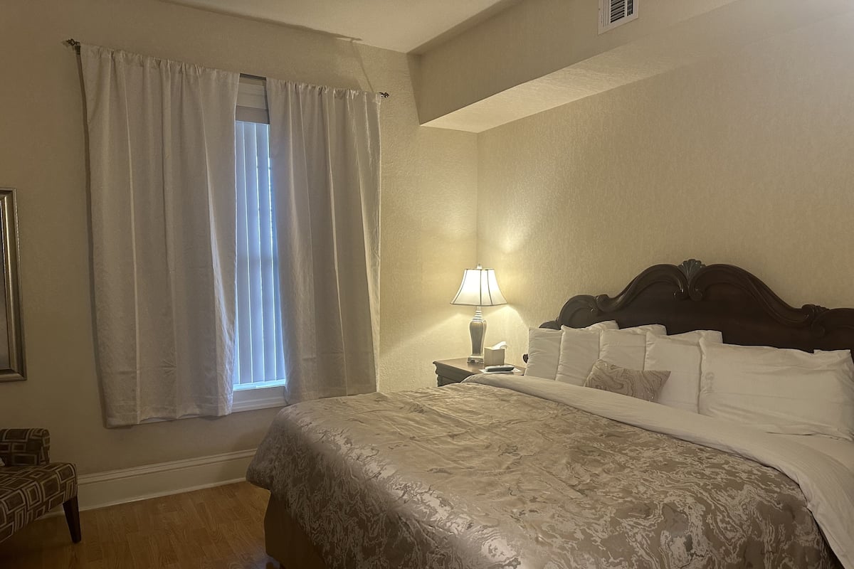 A hotel room with a large bed, a nightstand with a lamp, a window with curtains, and a patterned rug.