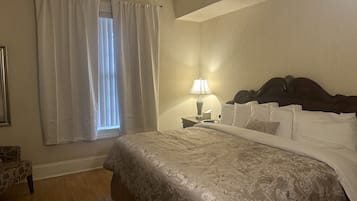 Un cuarto de hotel con una cama grande, una mesita de noche con lámpara, una ventana con cortinas y un tapete estampado.