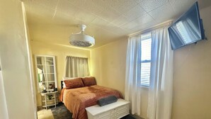 A bedroom with a bed, a dresser, a ceiling fan, a TV mounted on the wall, and a window with curtains.