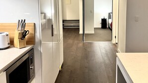 A modern kitchen with a microwave, toaster, and knife block. A hallway leads to a bedroom with a bed and a nightstand.