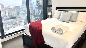 A hotel room with a large bed, a red throw blanket, and a view of a cityscape through a large window.