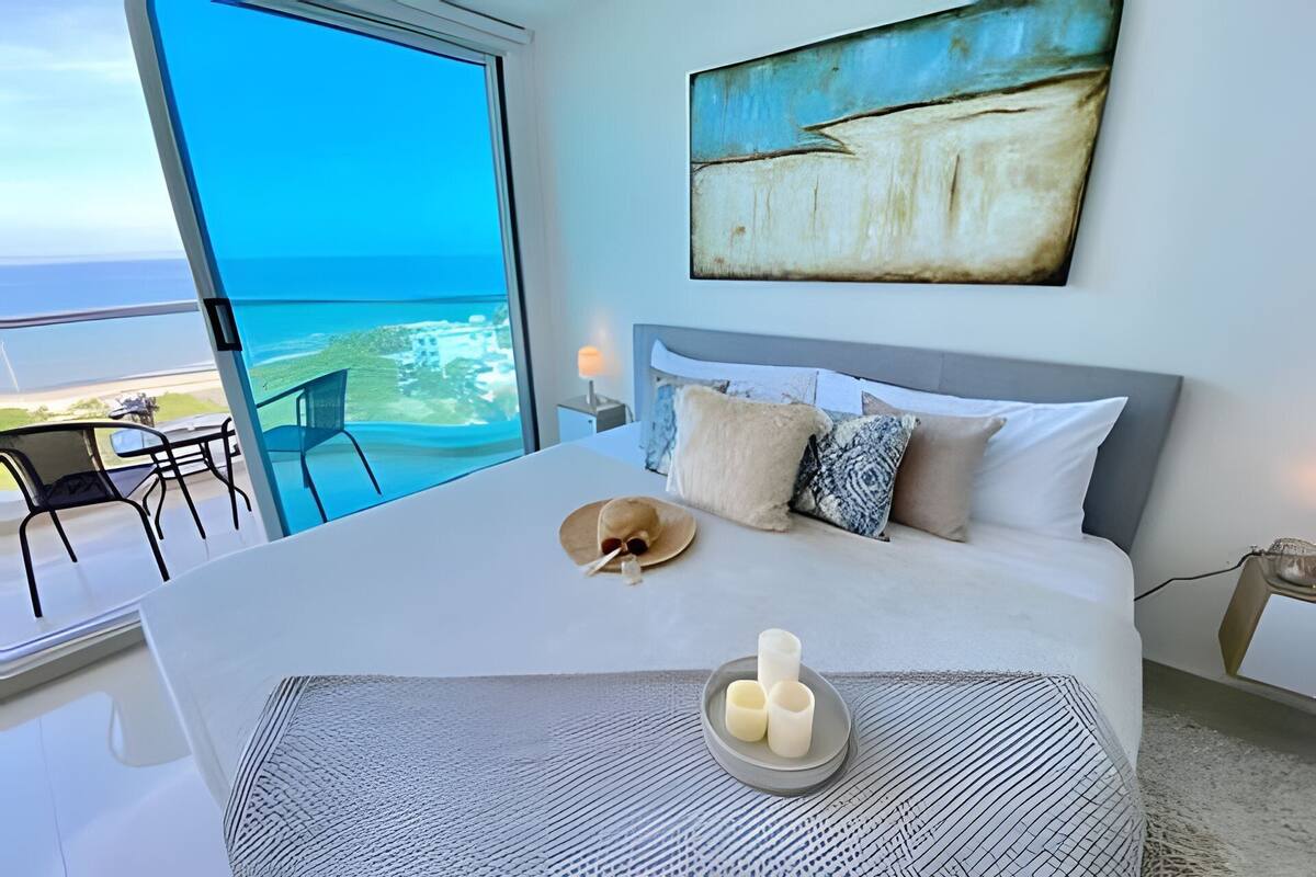 Une chambre avec un grand lit, une vue sur la mer, un tableau et un coin repas avec une chaise.
