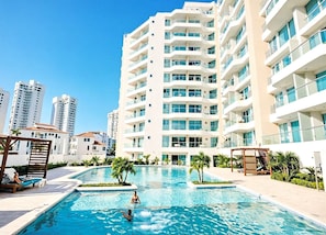 A modern hotel with a swimming pool, multiple balconies, and a view of surrounding high-rise buildings.