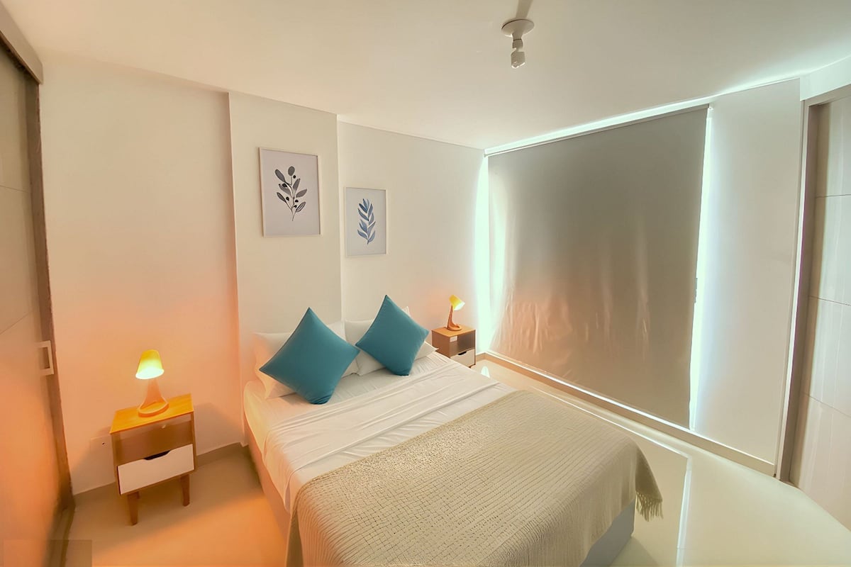 Une chambre à coucher comprenant un lit, deux oreillers, une table de chevet avec une lampe, un ventilateur de plafond et deux tableaux accrochés au mur.