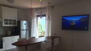A modern kitchen with white cabinets, a wooden dining table, and two bar stools. A large flat-screen TV is mounted on the wall displaying a scenic view.