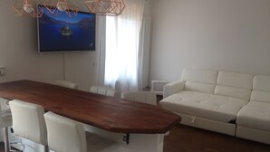 A modern living room with a wooden dining table, white leather sofa, and a flat-screen TV mounted on the wall.