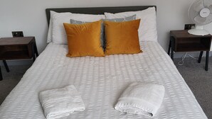 A neatly made bed with white bedding and two orange pillows, a bedside table with a lamp, and a wall-mounted fan. - Best Priced Apartment In Danum  19 (Doncaster)