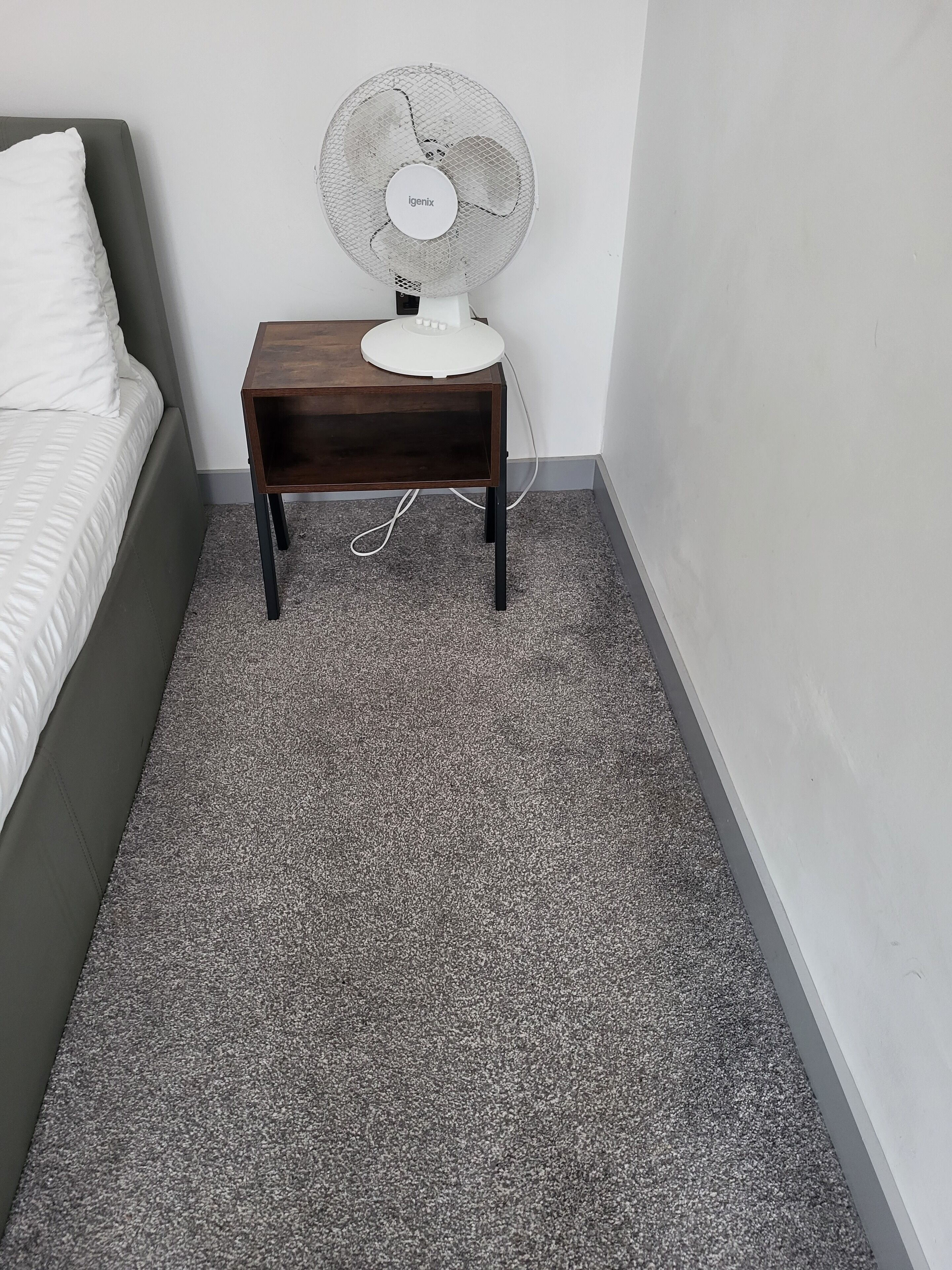 A bedroom with a bed, a small wooden nightstand, and a white fan.