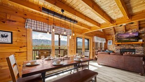 A wooden cabin interior with a dining table set for four, a fireplace, and a scenic mountain view through large windows.