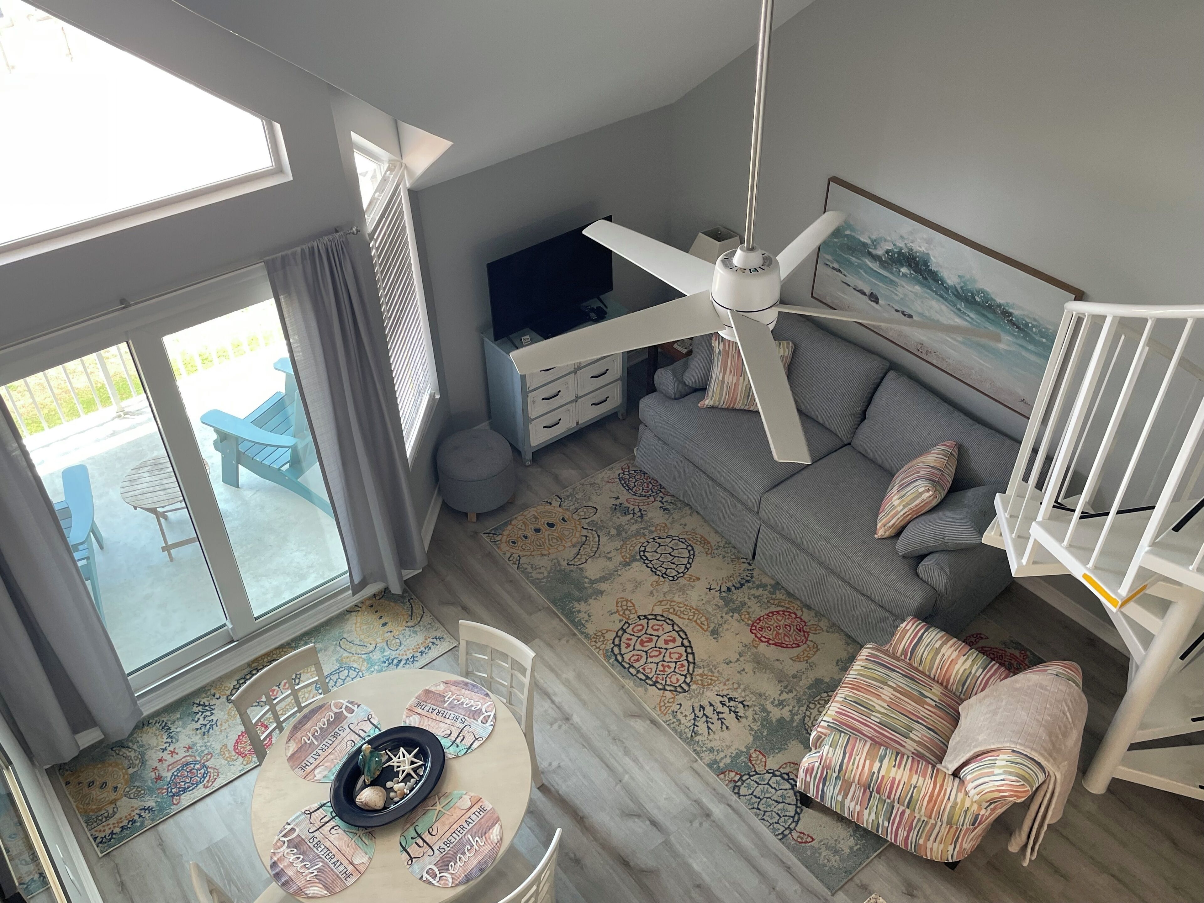 En rummelig stue med loftventilator, en grå sofa, et mønstret tæppe og et spisebord med strandtema-underlag.