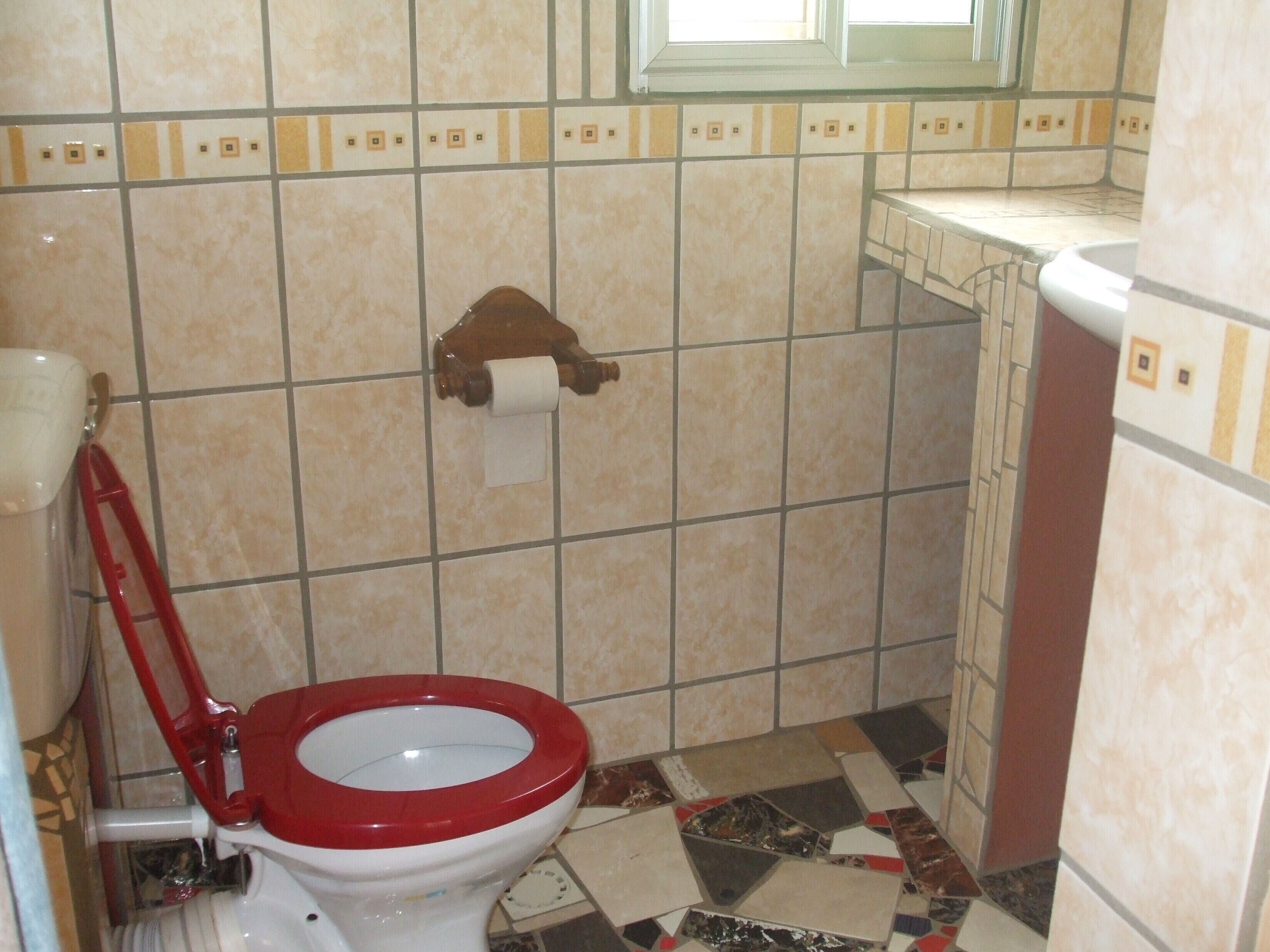 Une salle de bain avec des toilettes, un abattant de toilettes rouge, un porte-serviettes en papier et une fenêtre.