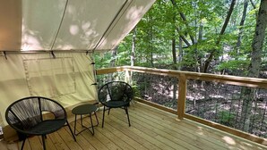 A covered deck with two chairs and a table, surrounded by a wooden railing, overlooking a forested area.