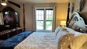 Une chambre avec un grand lit, une fenêtre avec des rideaux, une table de chevet avec une lampe et une commode avec un miroir.