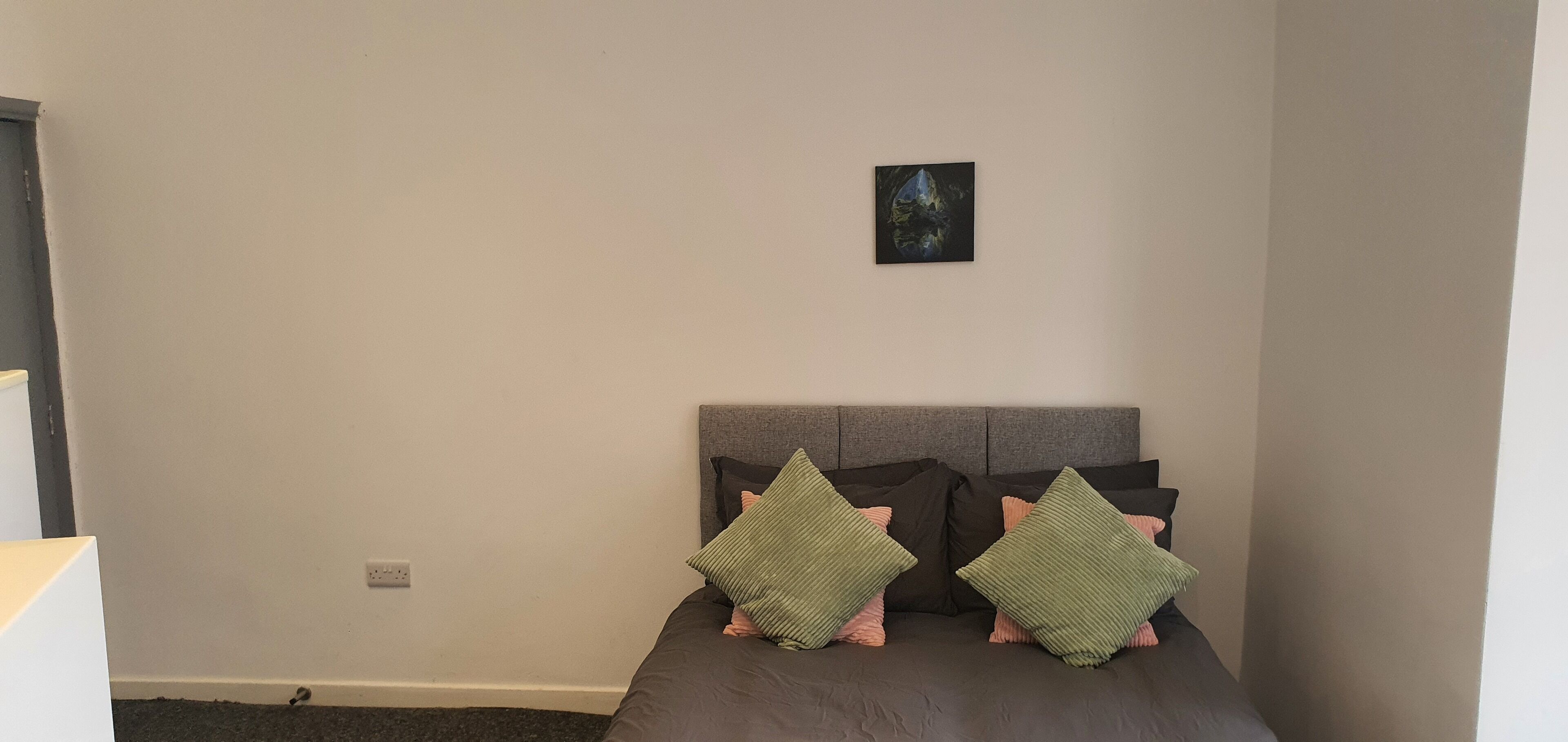 A bedroom with a grey headboard, a small framed picture on the wall, and two green pillows on the bed.