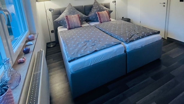 A modern bedroom with a large bed, two bedside lamps, a radiator, and a window with blinds. - Ferienwohnung "Am Landwehrgraben" (Michelau in Oberfranken)