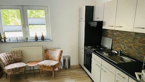 A compact kitchen with white cabinets, a black stove, and a window with plants. - Ferienwohnung "Am Landwehrgraben" (Michelau in Oberfranken)