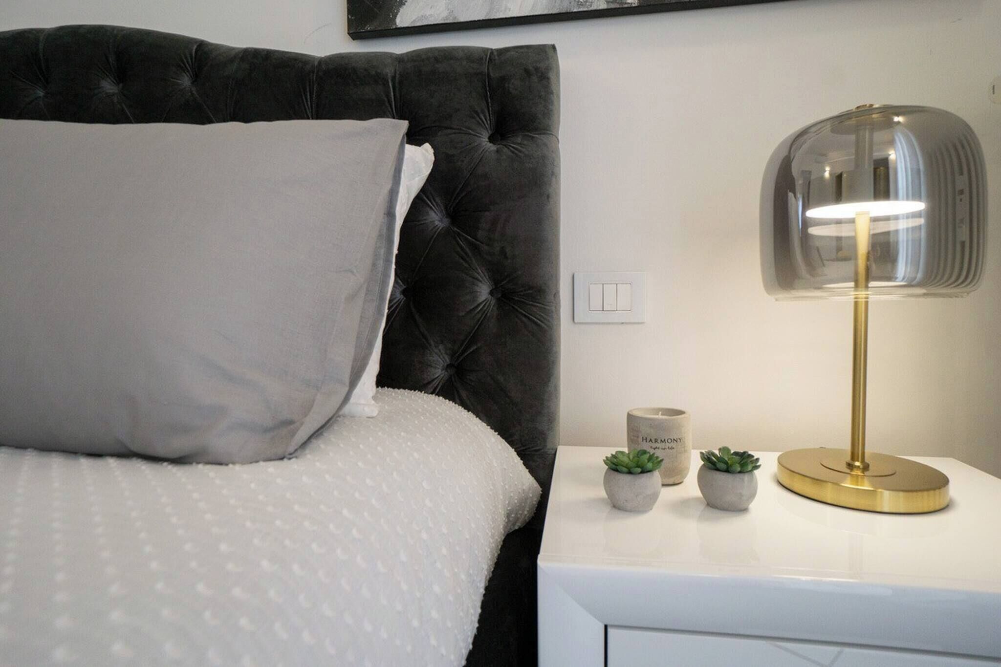 A bed with a tufted headboard, a white pillow, a white textured bedspread, a bedside table with a lamp, and two small potted plants.