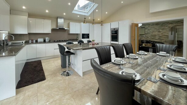 A modern kitchen with a central island, dining area with chairs, and a view of the city through skylights. - New Luxury Modern House Near Airport Free Parking (Luton)