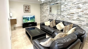 A modern living room with a black leather sofa, a glass coffee table, a flat-screen TV, a stone-textured wall, and a chandelier. - New Luxury Modern House Near Airport Free Parking (Luton)