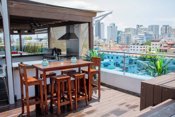 Un bar situé sur le toit d’un immeuble, avec une table et des chaises en bois, un barbecue et une vue sur la ville.