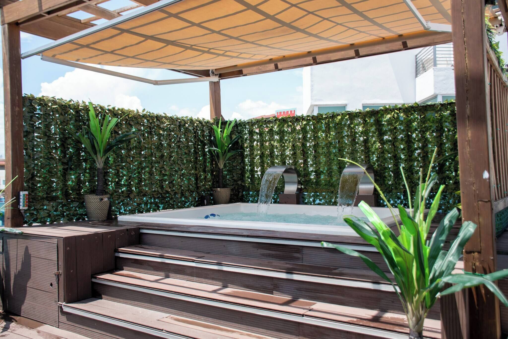 A hot tub with steps, surrounded by a wooden pergola and a hedge.