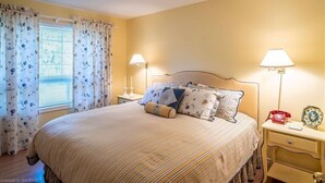 A bedroom with a large bed, floral curtains, two bedside lamps, and a nightstand with a telephone and decorative plate.
