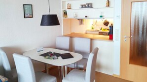 A compact room with a round table and four chairs, a kitchenette with a shelf above, and a door with a metallic handle. - Apartment Brockenblick in the Harz Mountains - Pool & Sauna (Braunlage)