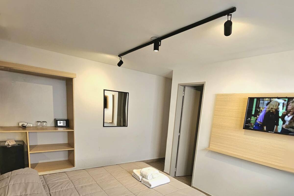 A modern hotel room with a large bed, a flat-screen TV mounted on the wall, a small desk with a computer, and a built-in shelf.