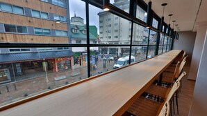 A modern bar with a long wooden counter and wicker chairs, large windows offering a view of the street and buildings outside. - M383 Hotel Bariloche (San Carlos de Bariloche)