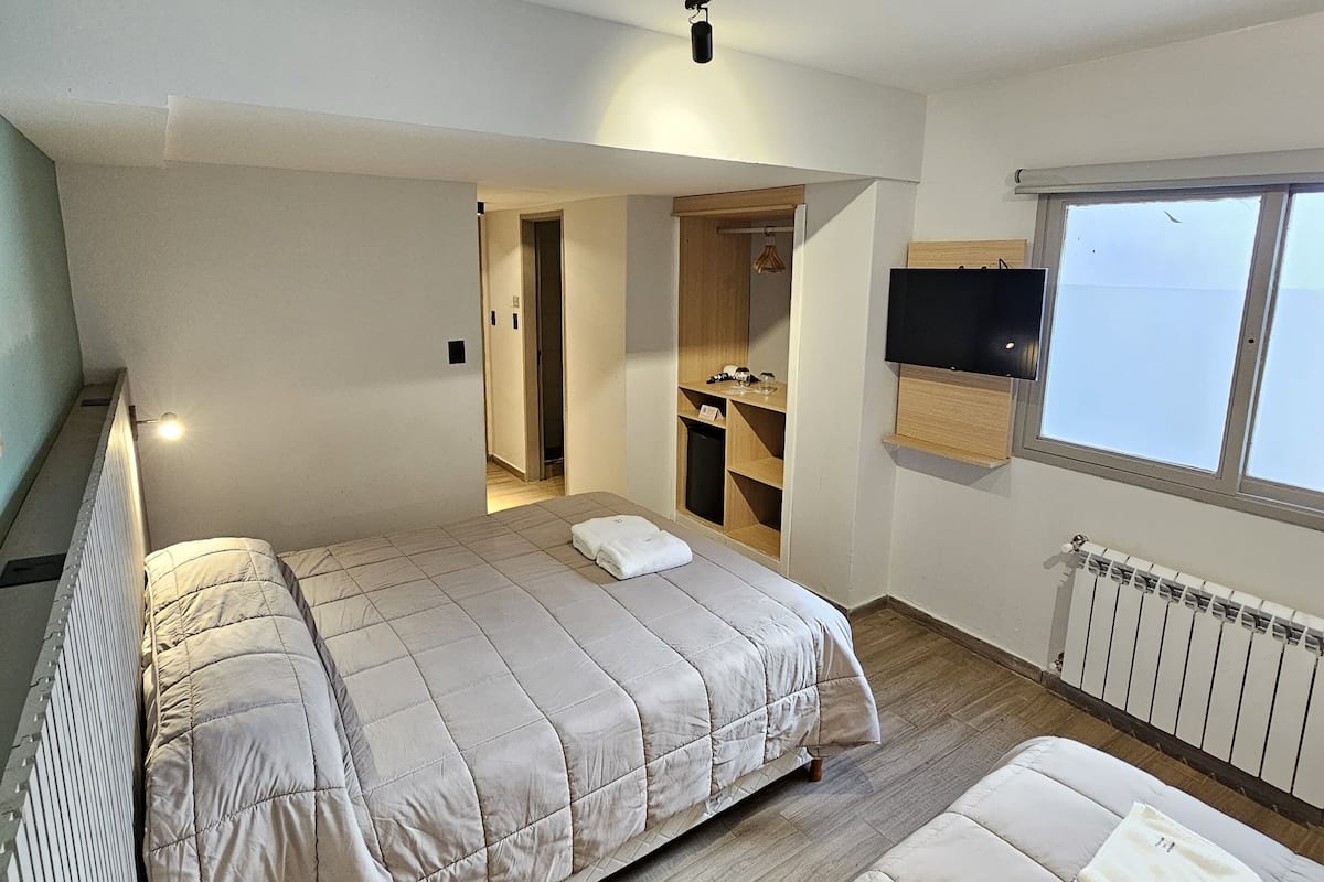 A modern bedroom with a large bed, a smaller sofa, a TV mounted on the wall, and a window providing natural light.
