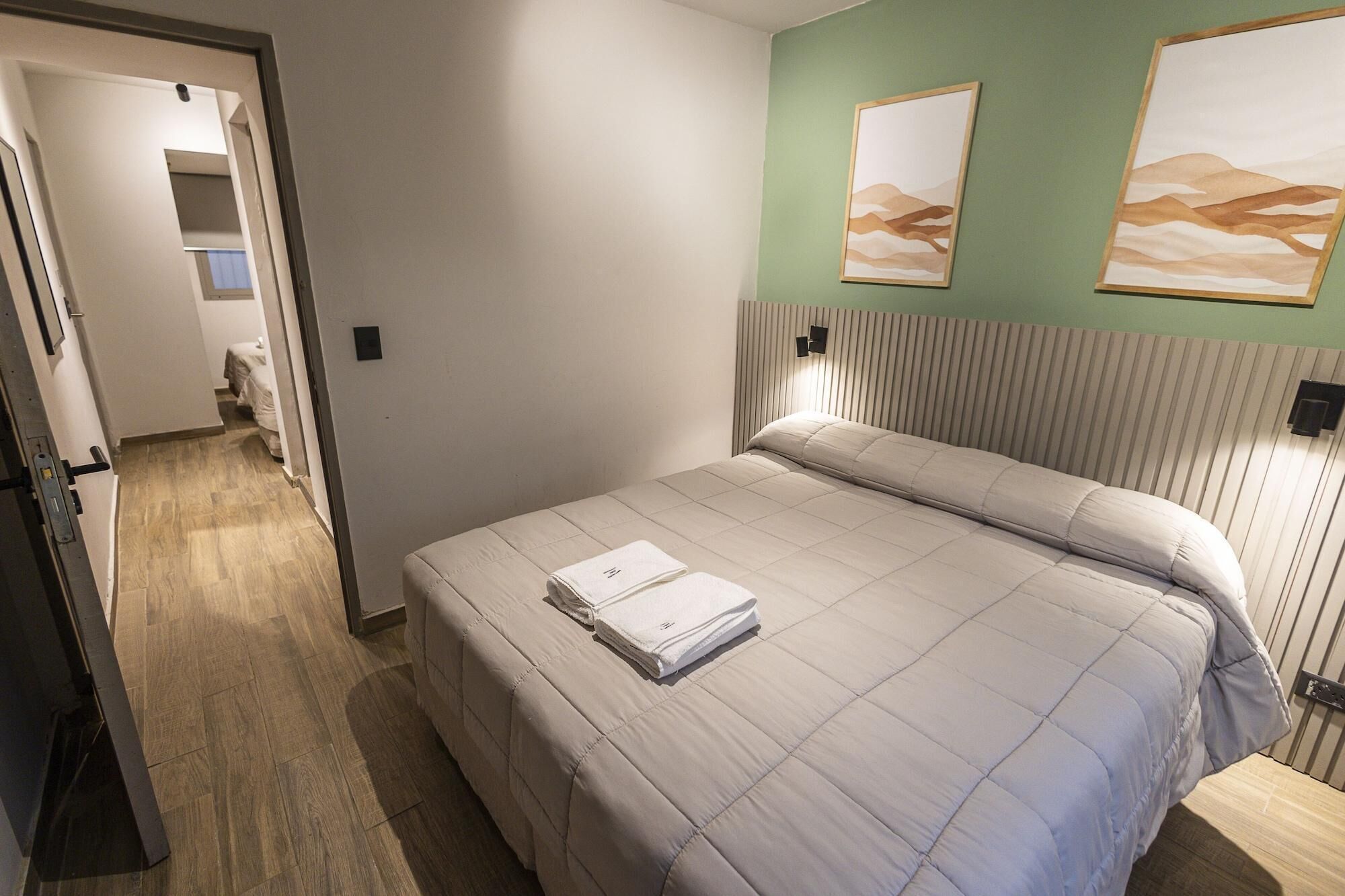 Una cama bien hecha con una toalla doblada, suelo de madera, pared verde y dos obras de arte enmarcadas.