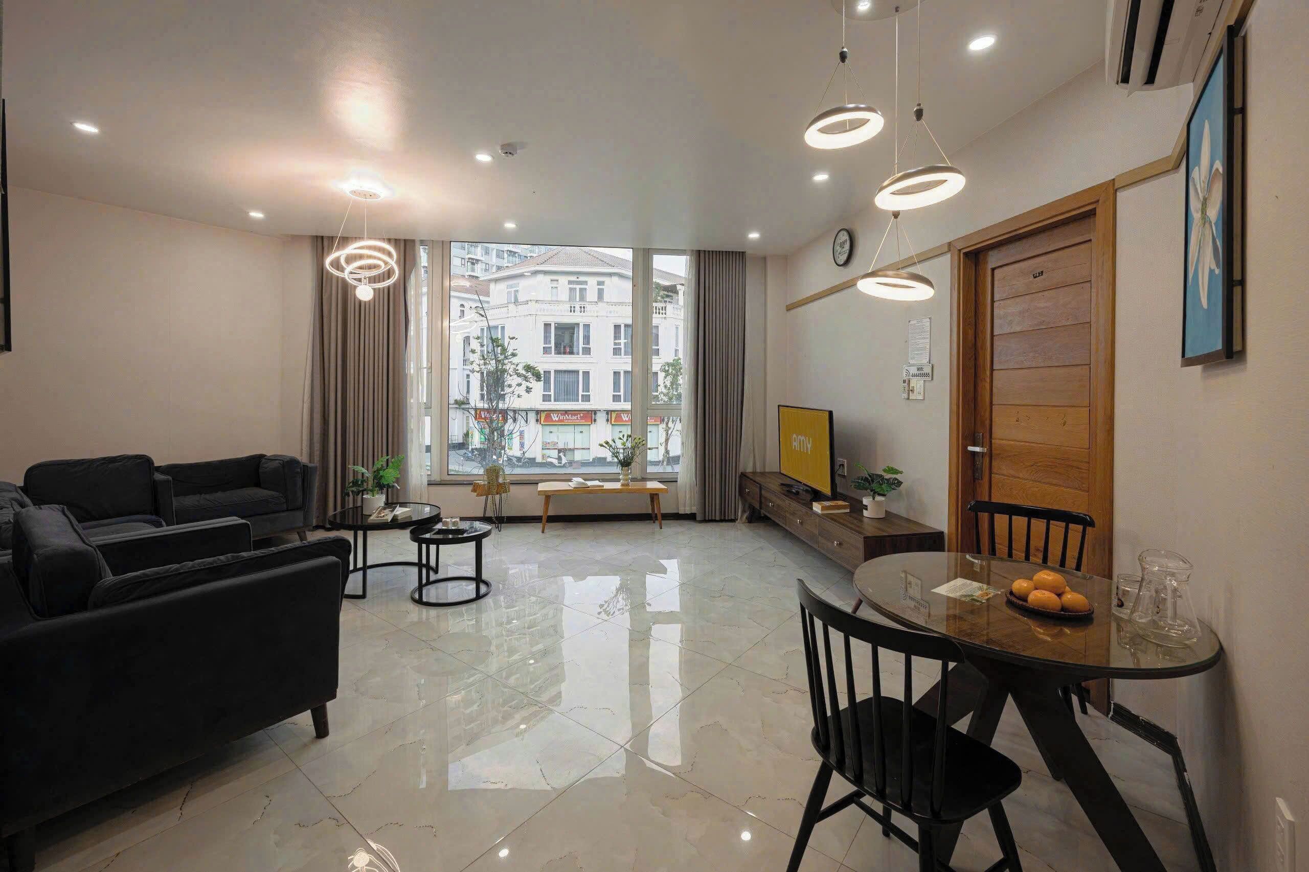 A modern living room with a black sofa, a round dining table with chairs, a TV, and a large window with a view of buildings.