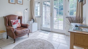 A cozy room with a brown leather armchair, a small white side table, a desk with a book, and a large window with a view of greenery.