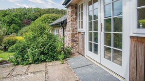 A stone building with large windows and a wooden door, surrounded by lush greenery.