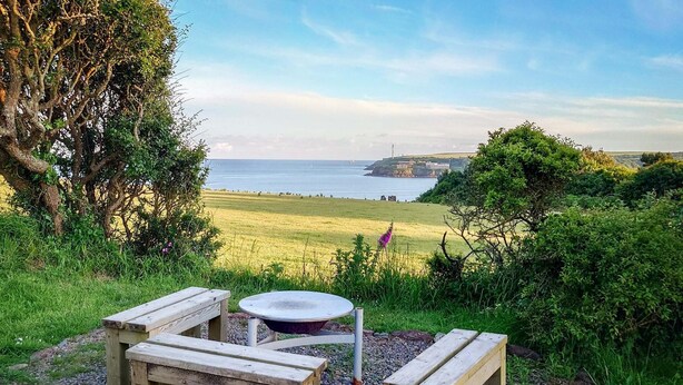 Un banc et une table en bois sont installés sur un chemin de gravier, offrant une vue sur la mer et une île lointaine.