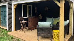 A covered outdoor seating area with a wooden bench, a wicker chair, and a yellow stool. - Binghamton's Beautiful Riverside Drive Living!
 (Binghamton)