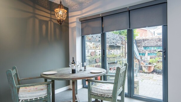 Comedor con una mesa redonda y cuatro sillas, una lámpara colgante, y vistas a un espacio exterior con un muro de ladrillo y plantas en macetas.