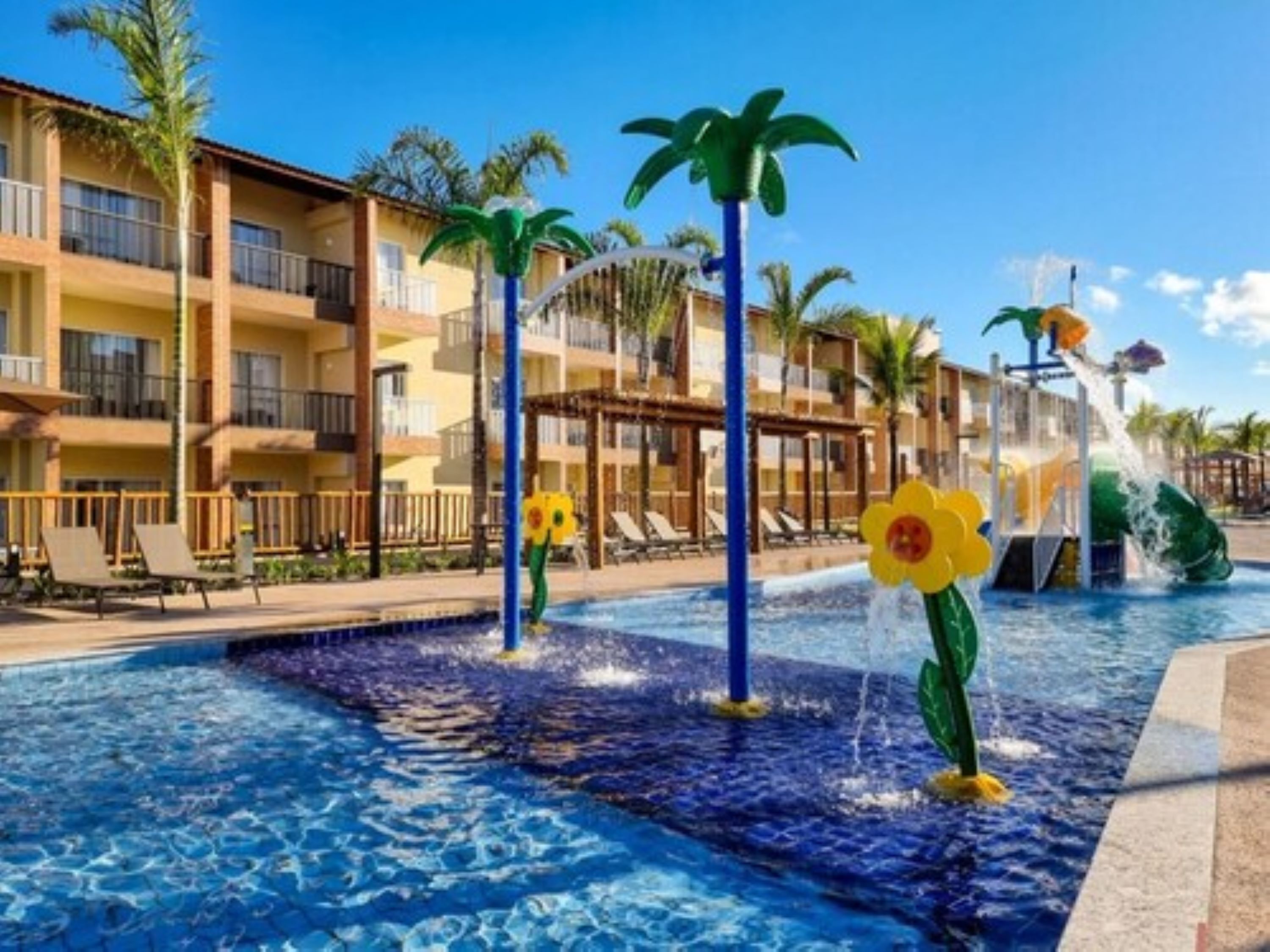 Zona de piscina de hotel con fuente de agua, palmeras y sillones de descanso.