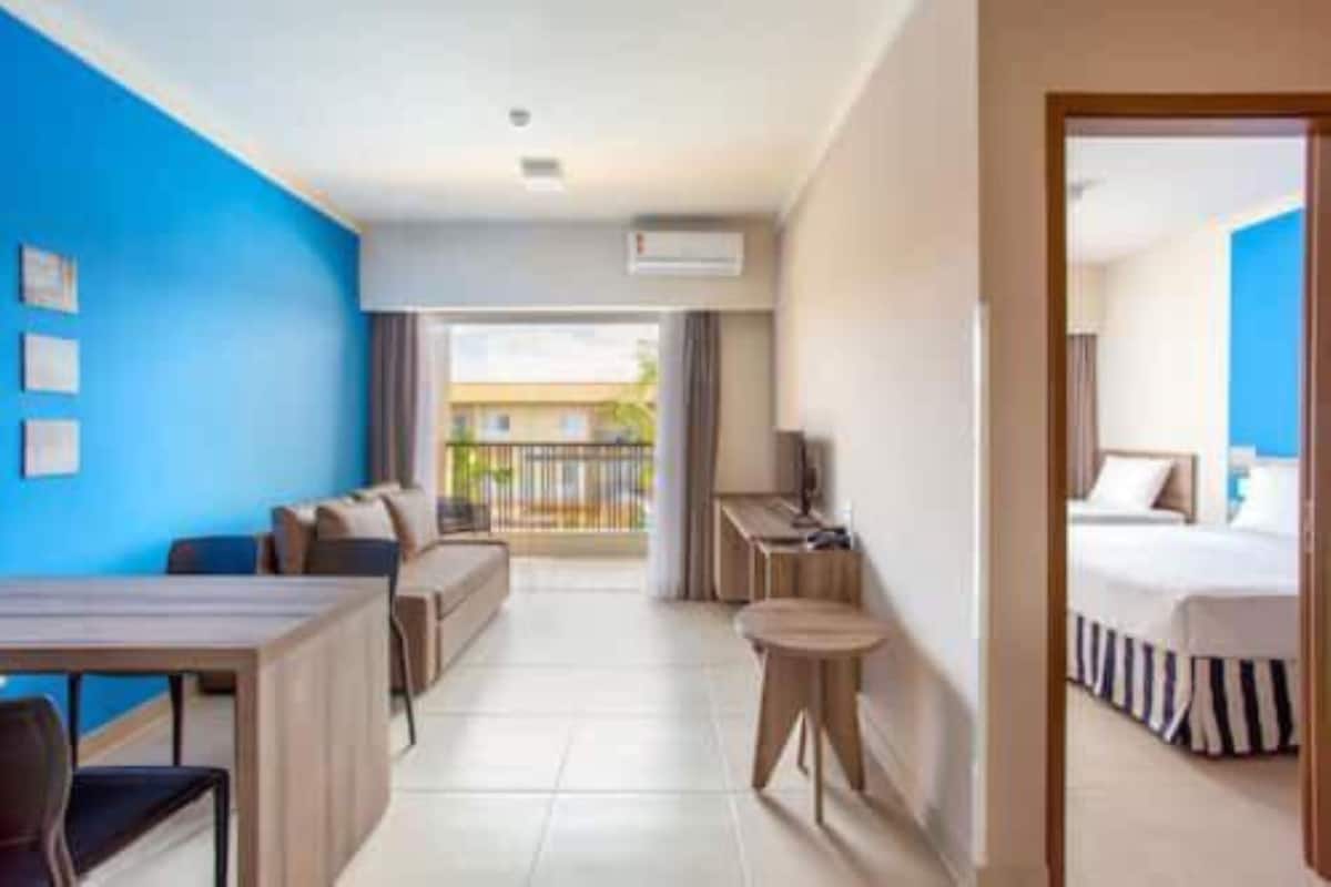 A hotel room with a bed, a desk, a chair, and a small table. There is a balcony with a view of the outside.